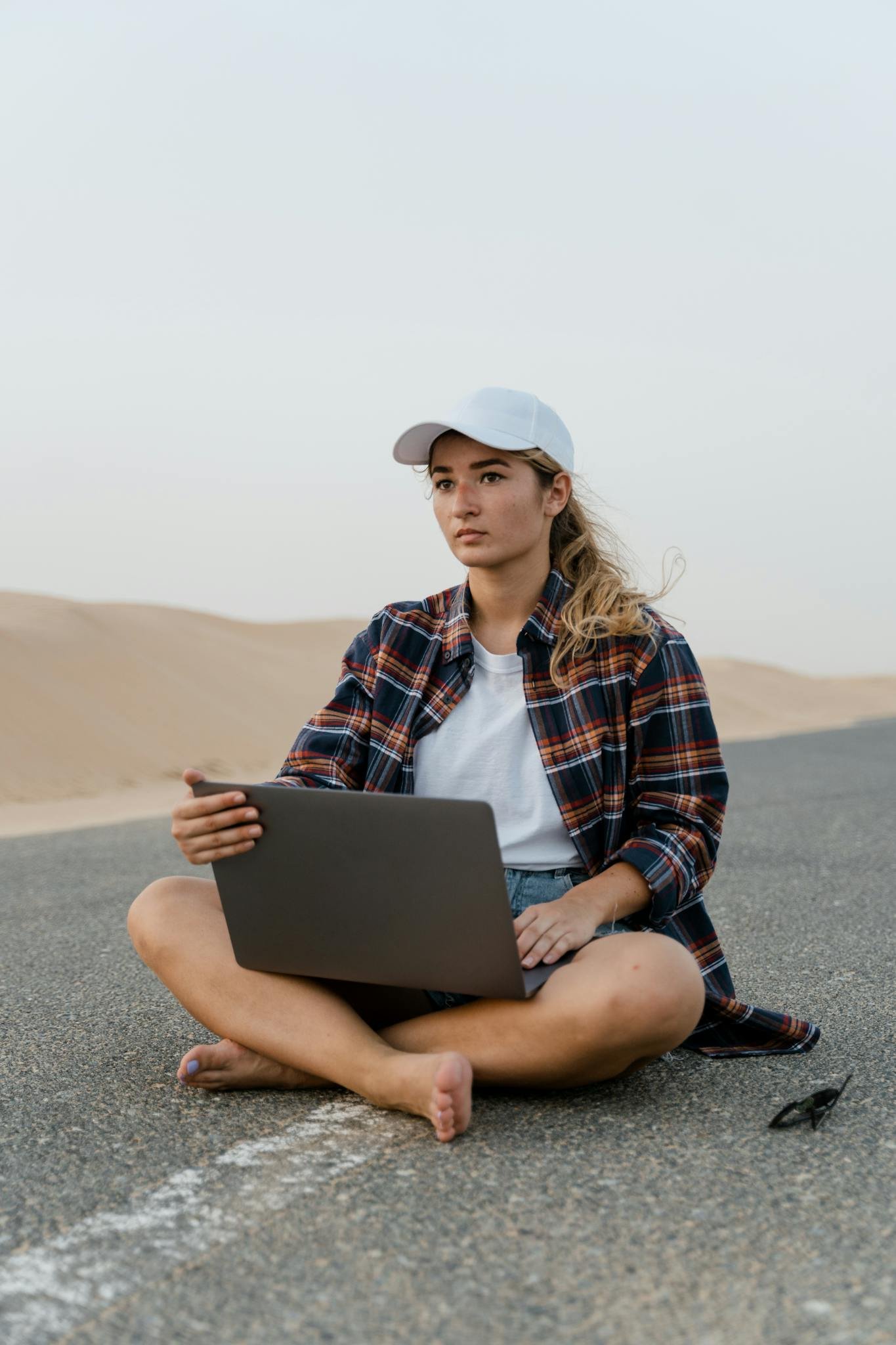 Young woman using laptop on a desert road, wearing casual attire and hat, symbolizing remote work.